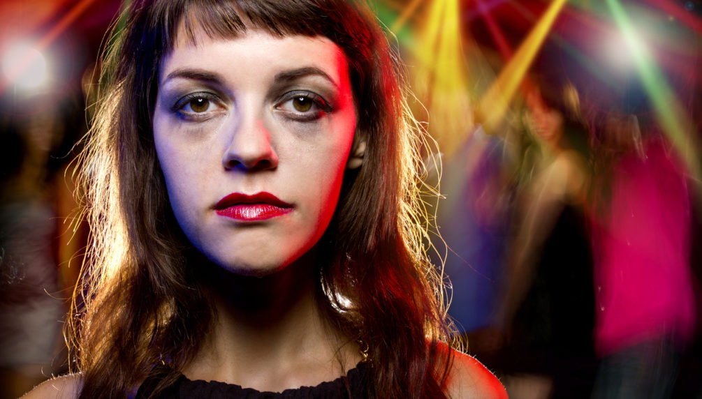 Disheveled drunk female with ruined make up in a nightclub. She is drunk or high on drugs. The people in the background are partying and dancing and motion blurred. The woman looks like an outcast and is self destructive by abusing drugs or alcohol.
