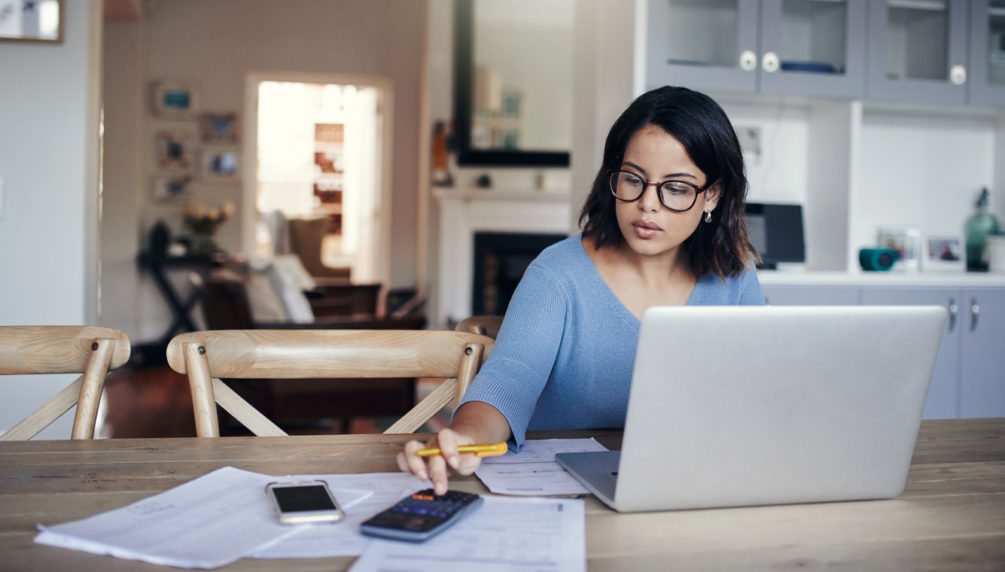 young woman using a laptop and calculator at kitchen table