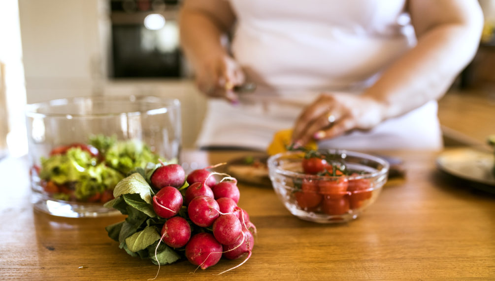 Unrecognizable overweight woman in white t-shirt at home preparing a delicious healthy vegetable salad in her kitchen.