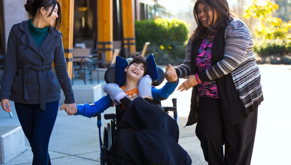 Eleven year old biracial disabled boy in wheelchair holding hands with caregivers while on a walk outdoors