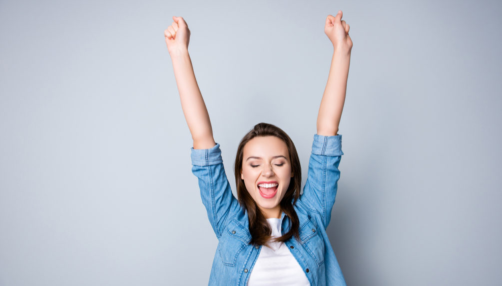 Amazed brunette young business woman in casual shirt is gesturing victory with her raised hands, she is shocked, extremely happy, with closed eyes, beaming smile, open mouth on grey background